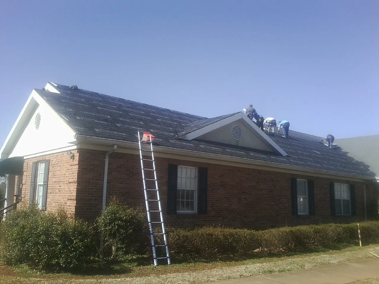 Crew tearing shingles off a brick ranch roof with a ladder set — Emergency Roof Repair work by our crew in Pall Mall