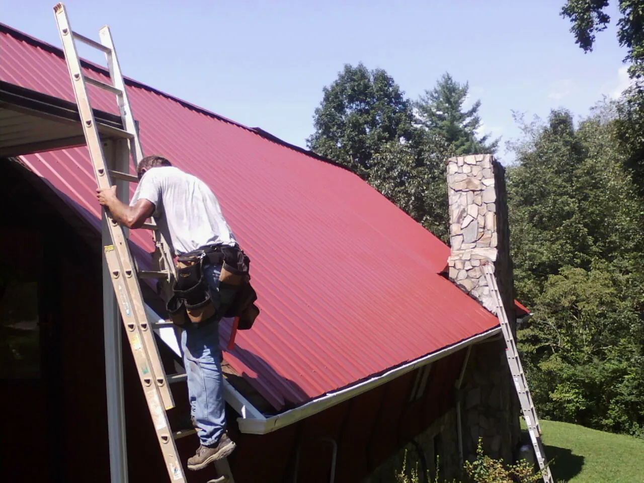 Roofer on a ladder beside a red metal roof and stone chimney — Soffit & Fascia Repair by Roofing Brothers in Pall Mall